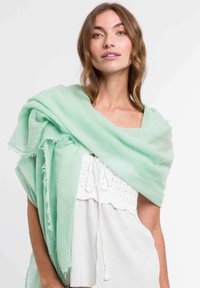 Șal de culoare verde deschis, țesut, care se așează pe umeri cu margini franjurate. Țesătura pare moale și ușoară, completând un top alb de dedesubt.