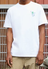 Henkilö, joka pitää päällään valkoista t-paitaa, jossa on pieni graafinen kuva juoksevasta maapallosta rinnassa, beigejä cargo-shortseja, seisoo ulkona.