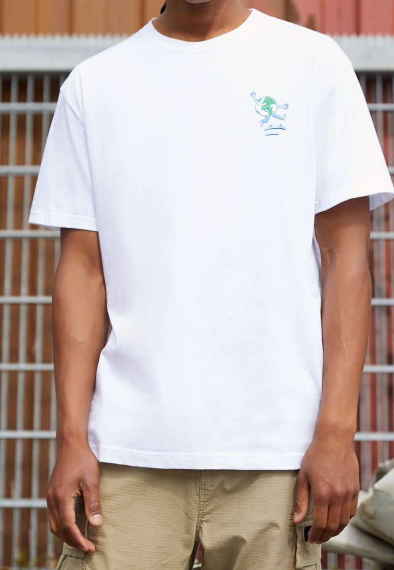 Henkilö, joka pitää päällään valkoista t-paitaa, jossa on pieni graafinen kuva juoksevasta maapallosta rinnassa, beigejä cargo-shortseja, seisoo ulkona.