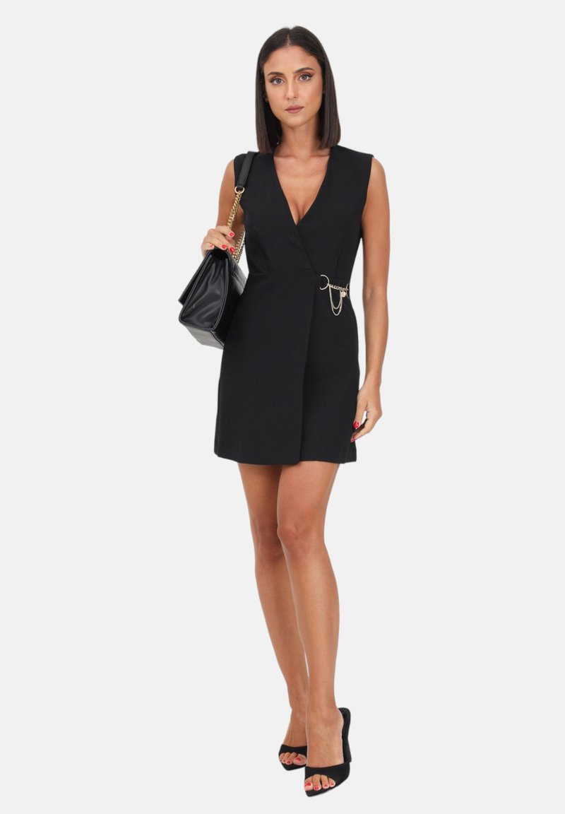 Schwarzes ärmelloses Kleid mit V-Ausschnitt, versehen mit einem goldenen Kettenakzent. Das Model hält eine schwarze Handtasche und trägt schwarze High Heels. Glattes Material.