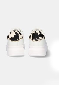 Zapatillas blancas con un talón de estampado de vaca en negro y blanco texturizado, con una parte superior lisa, suela acolchada y un acento dorado a lo largo del talón.
