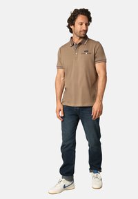 Homme debout portant un polo à manches courtes marron, un jean bleu foncé et des baskets blanches avec des accents bleus, regardant sur le côté.