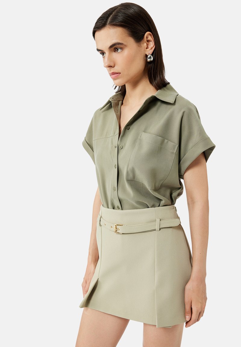 Femme aux cheveux foncés porte une chemise courte à boutons couleur olive et une mini-jupe beige ceinturée, debout devant un fond uni.