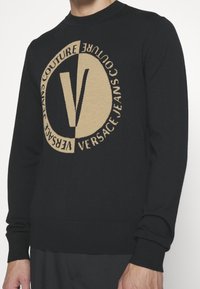 Pull en tricot noir avec un grand logo beige portant le texte "VERSACE JEANS COUTURE", poignets et ourlet côtelés.