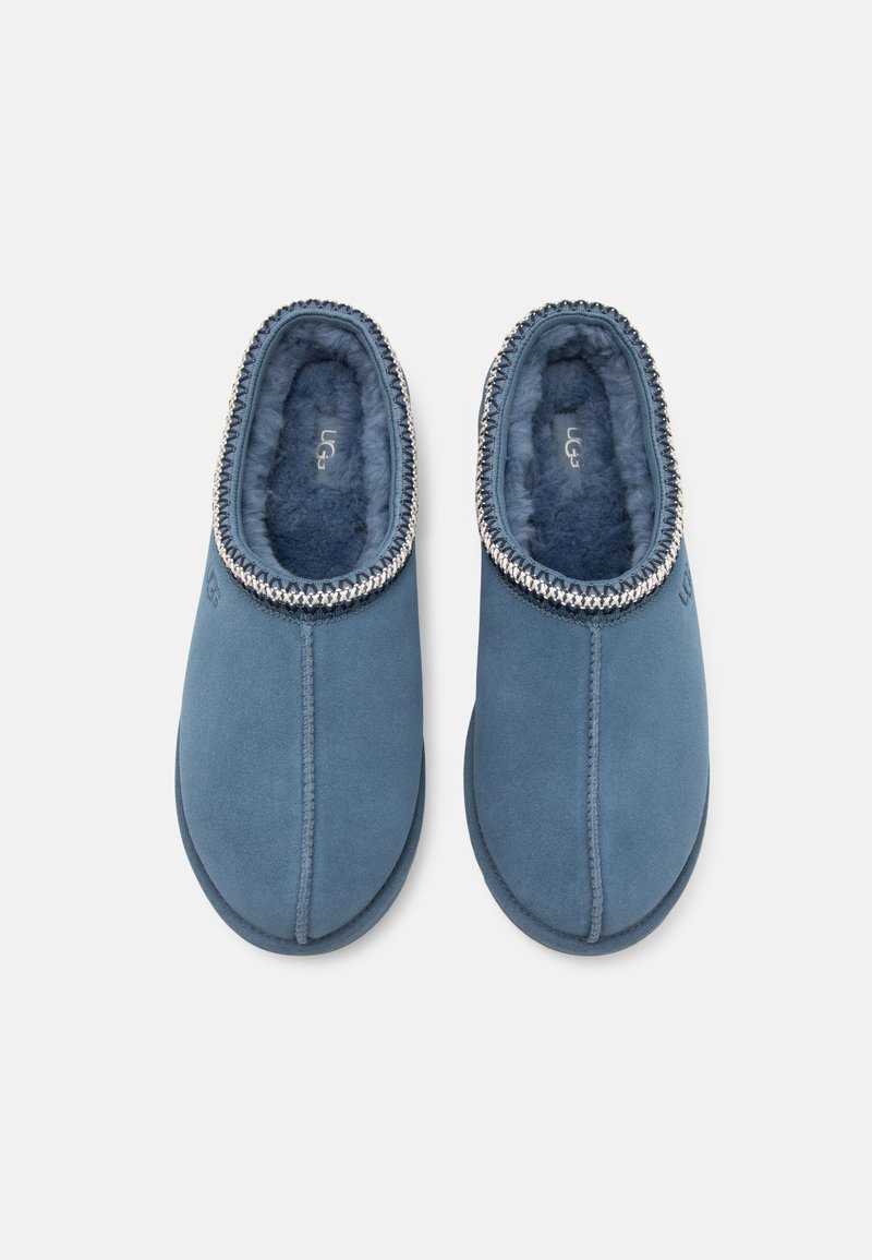 Pantoufles en suède bleu avec un intérieur doux et texturé, un design à bout rond et une bordure tressée autour de l'ouverture. Logo UGG visible.