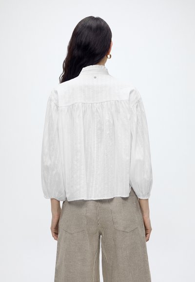 Mujer con cabello largo y oscuro que lleva una blusa blanca suelta con la espalda fruncida y pantalones anchos beige con un sutil patrón de rayas, vista desde atrás.