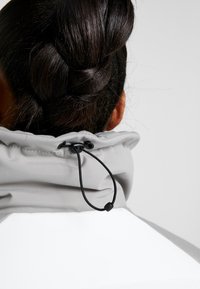 Grå jacka med krage och justerbar dragsnodd, med en mjuk textur och en vit panel längst ner. Håret är stylat i en flätad knut.