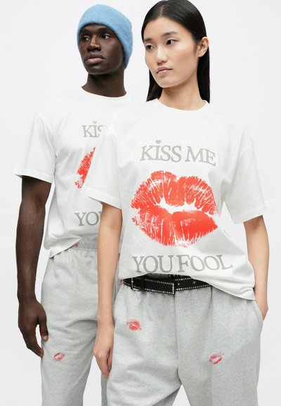 White cotton T-shirt featuring a red lip print and text "KISS ME YOU FOOL." Model wears gray sweatpants with scattered kiss prints and a black belt.