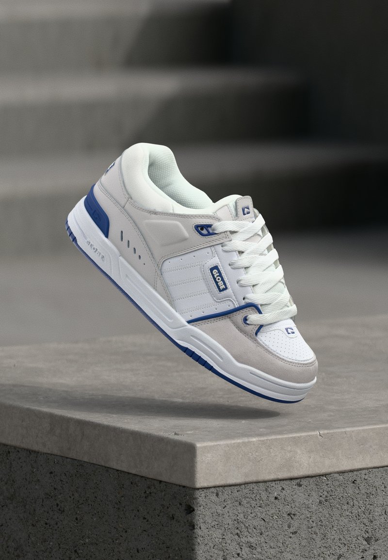 Baskets blanches et grises avec des accents bleus et des lacets, de la marque "Globe", flottant au-dessus d'une surface en béton avec des escaliers en arrière-plan.