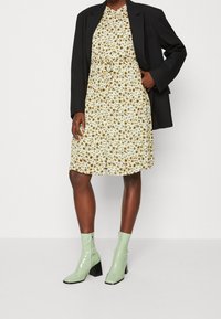 Abito stampato floreale in crema con fiori gialli e marroni, abbinato a un blazer nero e stivali alla caviglia verdi lucidi con tacco a blocco.