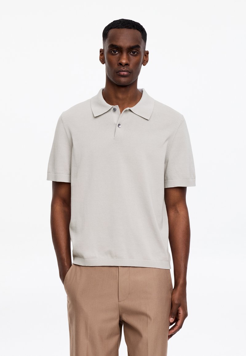 Homme à la peau foncée portant un polo à manches courtes beige clair et un pantalon marron clair, debout les mains dans les poches sur fond blanc.
