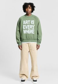 Felpa verde in cotone con il forte testo bianco "L'ARTE È OVUNQUE". Abbinata a pantaloni larghi beige e sneakers bianco e nero.