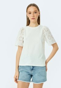 Hvid t-shirt med korte, broderede ærmer med bølgede kanter og blomsterudskæringer, parret med denim shorts. Glat bomuldsstruktur.