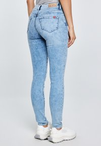 Naine seljas helesinised kitsad teksapüksid ja valged paksud tossud, seisab selg kaamerale, taust on lihtne.