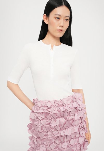 Top alb cu mâneci scurte și decolteu în stil henley, cu detalii cu nasturi, asociat cu o fustă roz cu volane, realizată dintr-un material satin, creând o textură în straturi.