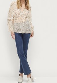 Blusa leggera e trasparente con motivo floreale beige, caratterizzata da maniche arricciate e scollatura con cordoncino, abbinata a jeans blu scuro e scarpe chiare.