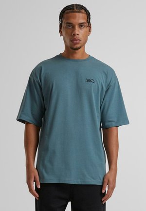 Tricou oversized de culoare teal, fabricat din bumbac, cu mâneci scurte, guler rotund și un mic logo negru pe partea stângă a pieptului.