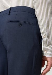 Dunkelblaue strukturierte Hose mit einer Gesäßtasche, die einen einzelnen Knopf hat. Kombiniert mit einem gestreiften hellen Hemd.