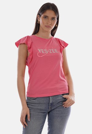 Donna che indossa un top rosa senza maniche con volant e la scritta "YES ZEE", abbinato a jeans blu, in piedi con una mano in tasca su sfondo bianco.
