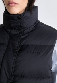 Svart puffer vest med høy krage, med en quiltet tekstur, trykknapper og en logo preget foran.