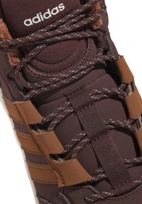 Brauner Sport-Sneaker aus strukturiertem Stoff, mit drei braunen Seitenstreifen, Schnürdesign und einer weichen braunen Innenauskleidung. Markenlogo sichtbar.