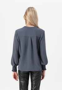 Grå bluse med smockede manchetter og plisseret ryg, med rund hals og tekstureret stof. Kombinert med svarte, blanke, tettsittende bukser.