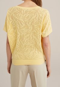 Pull en maille jaune avec un motif en vagues texturé, manches courtes et encolure arrondie. Le design comprend un ourlet côtelé et un matériau léger.