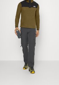 Sudadera de forro polar en verde oliva con canesú negro y cremallera de un cuarto, combinada con pantalones cargo gris oscuro con múltiples bolsillos. Sosteniendo una botella de agua gris.