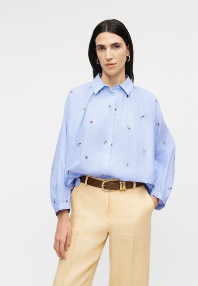 Γυναίκα που φοράει μπλούζα σε ανοιχτό γαλάζιο χρώμα με κουμπιά και μοτίβα μικρών κόκκινων λουλουδιών, μπεζ παντελόνι και καφέ ζώνη, στέκεται με το ένα χέρι στην τσέπη.