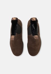 Zapatos de ante marrón tipo slip-on con paneles elásticos en los laterales, suela de goma plana y una etiqueta de cuero con la marca en el talón. Textura suave en general.