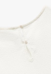 Hvid, tekstureret beklædning med rund halsudskæring og to lyse knapper bagpå. Blødt, fuzzy materiale med synlige sømme.