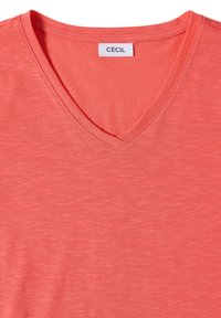 T-shirt rose corail à col en V avec une étiquette blanche indiquant "CECIL", cousue à l'intérieur de l'encolure.