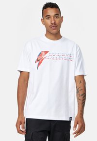 Hvid bomulds t-shirt med et David Bowie grafik, der har et rødt og blåt lynbolt design samt teksten "Aladdin Sane". Korte ærmer.