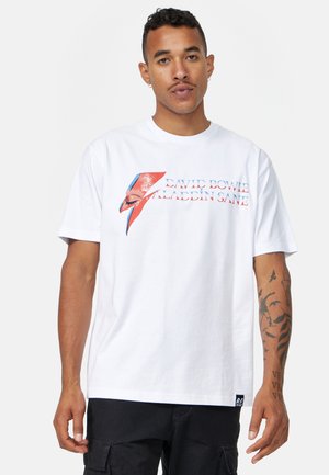 Vit bomull t-shirt med ett David Bowie-motiv som har en röd och blå blixtdesign samt texten "Aladdin Sane". Kortsleeve.