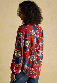 Rote Bluse mit langen Ärmeln im Blumendesign, mit blauen und cremefarbenen Blumen und Vögeln, aus weichem Stoff, lockerer Schnitt und mit gefalteten Rückendetails.