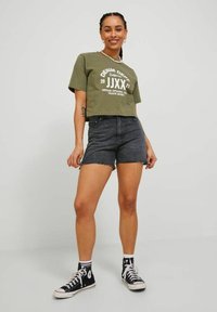 Camiseta corta color verde oliva con estampado blanco, combinada con shorts de mezclilla negros de tiro alto y zapatillas en blanco y negro.