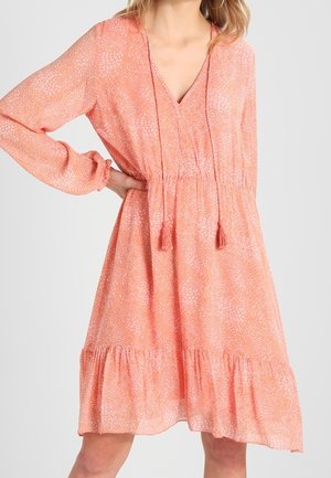 Femme portant une robe à manches longues corail avec encolure en V, cordon avec pompons, taille cintrée et ourlet à volants.