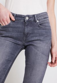 Mujer que lleva pantalones ajustados de mezclilla gris oscuro desgastados, con cierre de botón y cremallera, con una mano en el bolsillo, de pie contra un fondo liso.