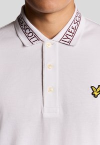 Witte polo shirt met een gebreide kraag, voorzien van een kastanjebruin geweven logo. Vier knopen bij de sluiting en een klein zwart en geel geborduurd vogel logo.