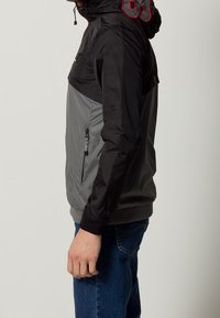 Huvjacka i svart och gråa material; har en dragkedjeficka, elastiska ärmslut och kontrasterande röd söm på huvan.