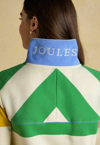 Frau von hinten trägt eine Jacke mit einem großen grünen und weißen Chevron-Muster und blauem Kragen, der mit "JOULES" bestickt ist.