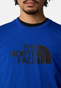 Camiseta de algodón azul con un gran logo negro. El cuello redondo y las mangas cortas complementan su diseño informal.