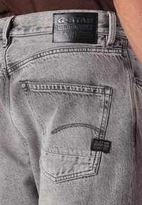 Jean en denim gris avec une taille haute. Présente une étiquette en cuir avec "G-Star Originals Raw Denim," des coutures contrastées et des détails de poches.