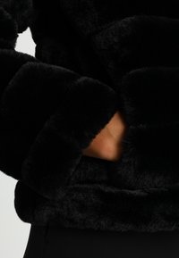 Veste en fausse fourrure noire avec une texture pelucheuse, une coupe décontractée et un design de poignets larges. Poche à main visible sur le côté gauche.