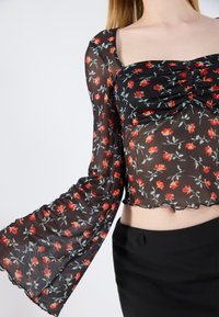Black floral crop top with long, flared sleeves. Sheer fabric featuring red flowers and green leaves. Gathered front detail and scalloped hem.