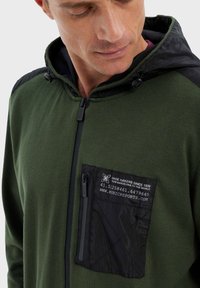 Veste zippée verte avec une capuche noire et des accentuations noires aux épaules. Elle possède une poche poitrine zippée noire avec un motif géométrique. Texture de matériau douce.