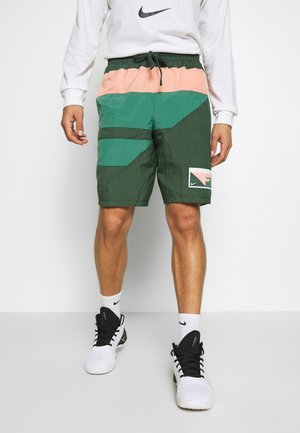 Mand iført farveblokkerede grønne og ferskenfarvede shorts, hvid Nike langærmet top, hvide Nike sokker og hvide og sorte sportssko.