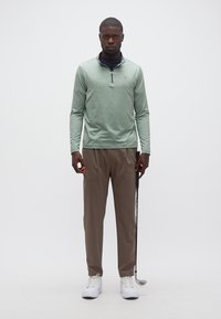 Άνδρας που στέκεται με γκολφ κλαμπ, φοράει πράσινο πουκάμισο με φερμουάρ μέχρι τη μέση, καφέ παντελόνι και λευκά αθλητικά παπούτσια, μπροστά σε απλό φόντο.