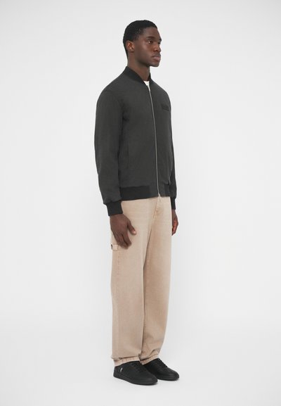 Μαύρο bomber μπουφάν με ριμπ μανσέτες και γιακά, εμπρόσθιο φερμουάρ, φορεμένο πάνω από λευκό πουκάμισο και συνδυασμένο με μπεζ παντελόνι και μαύρα αθλητικά παπούτσια.