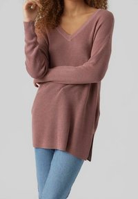 Femme portant un pull tricoté mauve à manches longues et col en V, ainsi qu'un jean bleu clair, debout les bras croisés devant un fond gris.